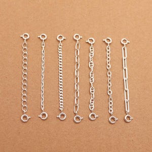 May include: Seven silver-colored chain extenders with different link styles are displayed against a brown background. Each extender has a clasp at both ends, designed to lengthen necklaces or bracelets. The chains vary in design, from delicate to more robust.