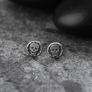 Op de afbeelding: Twee zilverkleurige oorstekers. Elke oorbel heeft een schedelontwerp in een cirkelvormige lijst met een getextureerde rand. De oorbellen liggen op een grijze, getextureerde ondergrond, wat het glanzende metaal accentueert.