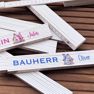 Könnte beinhalten: Weiße Zollstöcke mit schwarzen Markierungen und personalisiertem Text. Die Zollstöcke liegen auf einer Holzoberfläche. Einige Zollstöcke haben Namen und Hausillustrationen in Rosa und Blau.