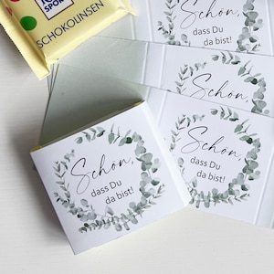 Könnte beinhalten: Zwei Ritter Sport Schokoladentafeln, eine gelbe mit Schokolinsen und eine gelbe mit Weisse Zitrone, sowie drei weiße rechteckige Karten mit einem grünen Eukalyptuskranz-Motiv und dem Text Schön, dass Du da bist!
