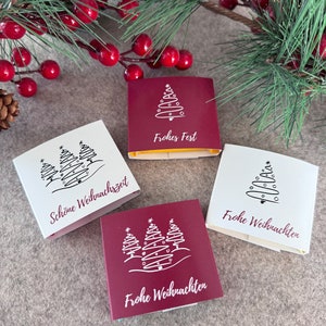 Pode incluir: Quatro pequenas caixas de presente retangulares com tema natalino. Duas s&atilde;o brancas e duas s&atilde;o bord&ocirc;. Cada caixa apresenta uma ilustra&ccedil;&atilde;o branca de &aacute;rvores de Natal e texto em alem&atilde;o. As caixas est&atilde;o dispostas sobre uma superf&iacute;cie de cor neutra, com vegeta&ccedil;&atilde;o festiva ao fundo.