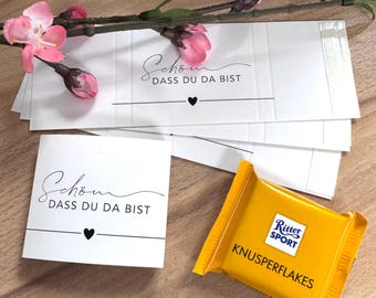 Op de afbeelding: Witte kaarten met de tekst "Schön dass du da bist" en een hartsymbool, naast een gele Ritter Sport chocoladereep. Roze bloemen en knoppen zijn gerangschikt op een houten oppervlak.