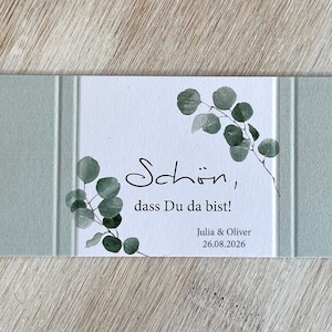 May include: A white card with green eucalyptus leaves printed on it. The card says "Sch&ouml;n, dass Du da bist!" and "Julia & Oliver 26.08.2026". The card is surrounded by a light green border.