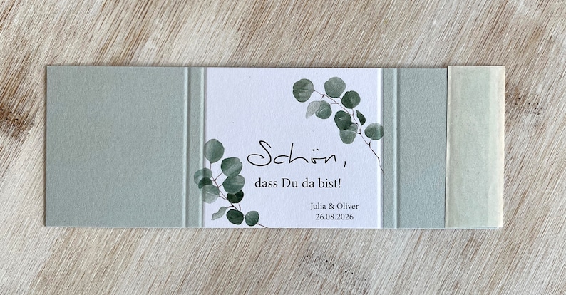 May include: A white card with green eucalyptus leaves printed on it. The card says "Sch&ouml;n, dass Du da bist!" and "Julia & Oliver 26.08.2026". The card is surrounded by a light green border.