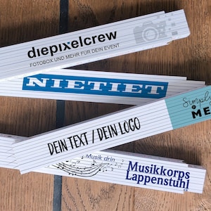 Könnte beinhalten: Mehrere weiße Zollstöcke mit schwarzem Text und blauen Akzenten liegen auf einer Holzoberfläche. Die Zollstöcke zeigen Sätze wie "diepixelcrew" und "DEIN TEXT / DEIN LOGO". Ein Zollstock hat den Text "Simply ME".