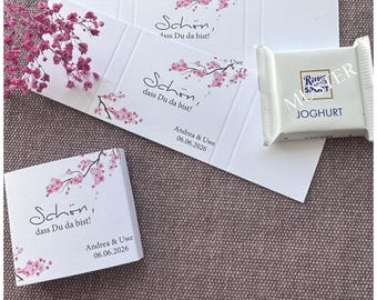 Pode incluir: Embalagens de papel branco e uma barra de chocolate quadrada com um rótulo Ritter Sport Joghurt. As embalagens apresentam ilustrações de flores de cerejeira cor-de-rosa, o texto "Schön, dass Du da bist!" e os nomes "Andrea & Uwe" com a data "06.06.2026".