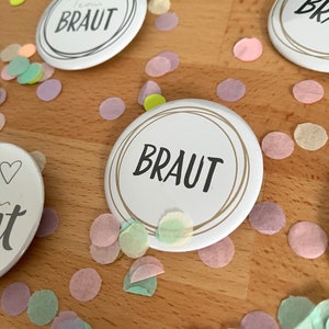 Könnte beinhalten: Zwei weiße runde Knöpfe mit goldener Umrandung und schwarzem Text. Der Text auf den Knöpfen lautet "Braut". Die Knöpfe sind von pastellrosa, grünen und gelben Konfetti umgeben.