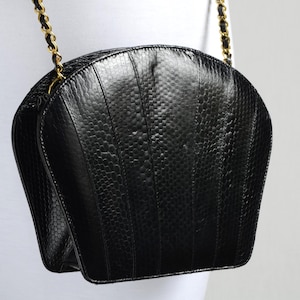 May include: Black handbag with a textured finish and a gold chain strap. The bag has a distinctive, fan-shaped design and a subtle, reptile-like pattern. The chain is gold.