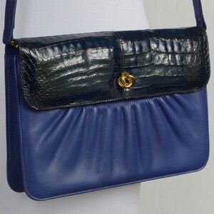 May include: A blue leather crossbody bag with a textured, dark blue flap and a gold-tone clasp. The bag has a gathered design on the front and a matching blue strap.