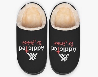 May include: Black house slippers with a cream-colored faux fur lining. The slippers have the words "Addicted to Jesus" printed on them in white and red, along with a cross graphic.