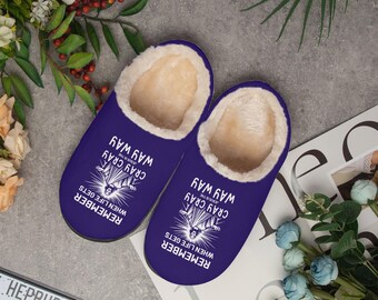 May include: A pair of purple slippers with a white sherpa lining. The slippers have the text "REMEMBER WHEN LIFE GETS CRAY CRAY WAY WAY" printed in white. The slippers are on a gray surface with flowers.