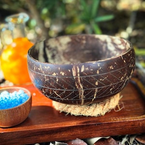 Puede incluir: Un cuenco de coco marrón oscuro con patrones geométricos grabados se encuentra sobre una pequeña esterilla tejida. El cuenco está sobre una bandeja de madera, con un pequeño cuenco de madera con cristales azules y una botella de líquido naranja en el fondo.