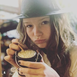 May include: A young person wearing a black fedora hat holds a small black cauldron with a golden liquid bubbling inside. The cauldron has a handle and a small wooden stick is stirring the liquid.