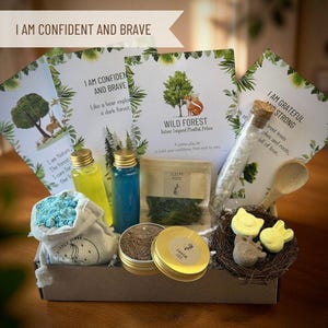 May include: A gift box with various items, including bottles of liquid, a small bag of blue crystals, and small tins. The box also contains cards with text that reads "I AM CONFIDENT AND BRAVE" and "WILD FOREST". A wooden spoon and bird-shaped soaps are also included.