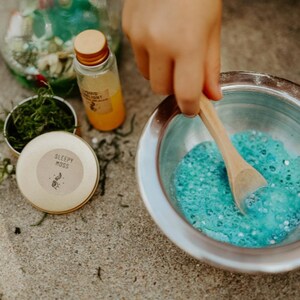 May include: A child's hand stirs blue, bubbly liquid in a silver bowl with a wooden spoon. A small bottle of orange liquid and a container labeled "Sleepy Moss" are nearby, suggesting a craft or sensory activity.