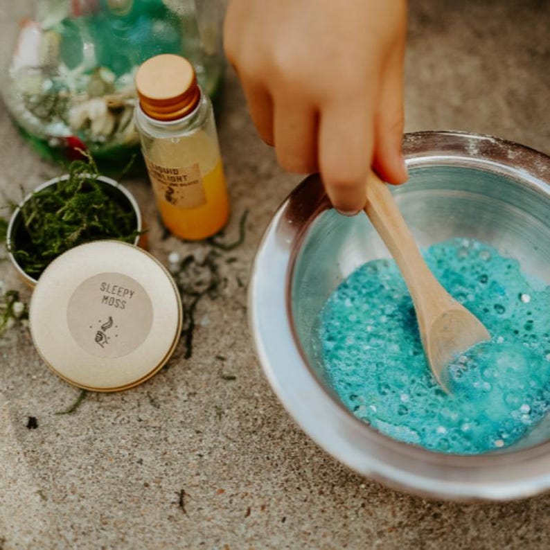 May include: A child's hand stirs blue, bubbly liquid in a silver bowl with a wooden spoon. A small bottle of orange liquid and a container labeled "Sleepy Moss" are nearby, suggesting a craft or sensory activity.