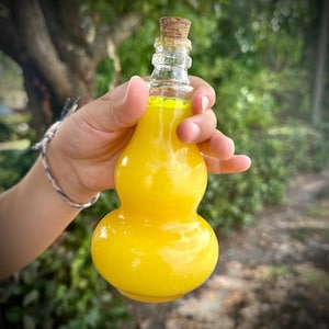 May include: A clear glass bottle with a gourd-like shape, filled with bright yellow liquid. The bottle has a cork stopper and is held in a hand. The background is blurred, with green foliage visible.