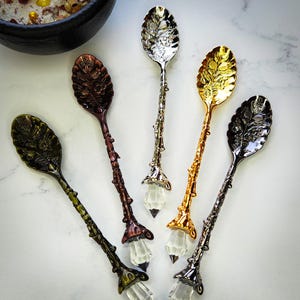 May include: An assortment of decorative spoons with ornate leaf-shaped bowls and crystal-like accents. The spoons come in various metallic finishes, including gold, silver, bronze, and black, and are arranged on a white surface. The spoons are likely for serving or display.