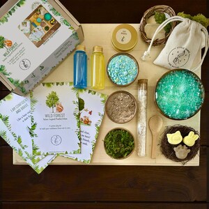 May include: A Wild Forest sensory play kit with natural ingredients. Includes a box, bottles, bowls of colorful materials, a nest with soap, a wooden spoon, and cards with text. The kit is designed to empower children through mindful play.