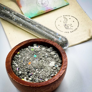 May include: A wooden bowl filled with gray sand and iridescent glitter. A glass test tube with gray sand and iridescent glitter is next to the bowl. The text "Little Hands and Nature" is on a white bag behind the bowl.
