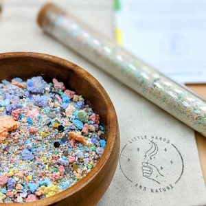 May include: A wooden bowl filled with colorful, textured materials, including iridescent pieces and small, irregular shapes. A clear tube filled with similar materials is in the background, along with a canvas bag with the text "LITTLE HANDS AND NATURE".