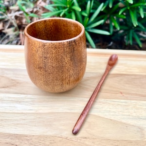 May include: A wooden cup and spoon set. The cup is brown and has a rounded shape. The spoon is long and slender, also made of wood.