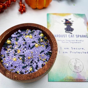 May include: A wooden bowl filled with purple, black, gold, and white sprinkles. The bowl is sitting on a white surface next to a card that reads "Stardust Cat Sparkle" and "Nature Inspired Mindful Potion Ingredient". The card also says "I am Secure, I am Protected" and has a small illustration of a black cat wearing a witch's hat.