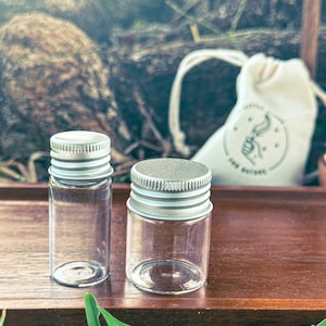 May include: Two small, clear glass bottles with silver metal screw-on lids. A small, white drawstring bag with a printed design is in the background. The bottles are on a dark wooden surface.