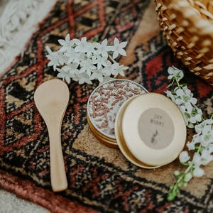 May include: A wooden spoon rests beside an open, round, gold-colored tin filled with small, brown and white sprinkles. The tin's lid, also gold, has a label that reads "Dry Worms." White flowers and a woven basket are in the background, all on a patterned rug.