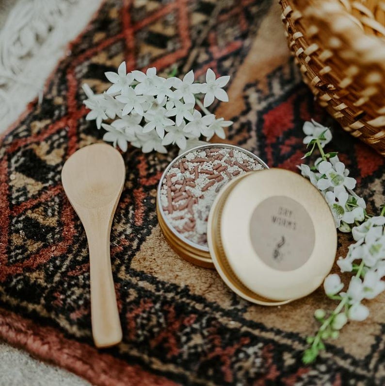 May include: A wooden spoon rests beside an open, round, gold-colored tin filled with small, brown and white sprinkles. The tin's lid, also gold, has a label that reads "Dry Worms." White flowers and a woven basket are in the background, all on a patterned rug.