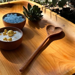 May include: A wooden heart-shaped spoon rests on a wooden tray, next to two small wooden bowls filled with blue crystals and white granules with gold spheres. A succulent plant is also on the tray.