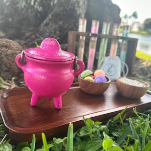 May include: A bright pink miniature cauldron with a lid and three legs sits on a wooden tray. Small wooden bowls hold colorful items. Test tubes and a small bag are in the background, suggesting a mystical or apothecary theme.
