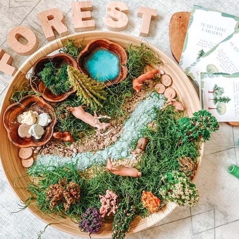 May include: A wooden tray with a miniature forest scene, including a blue pond, river, and various plants. Small animal figurines and wooden accents are scattered throughout the scene. The word "FOREST" is spelled out in wooden letters.