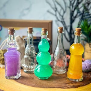 May include: Five glass bottles with cork stoppers filled with different colored liquids. The bottles are arranged in a row on a wooden surface. The liquids are purple, green, clear, and yellow.