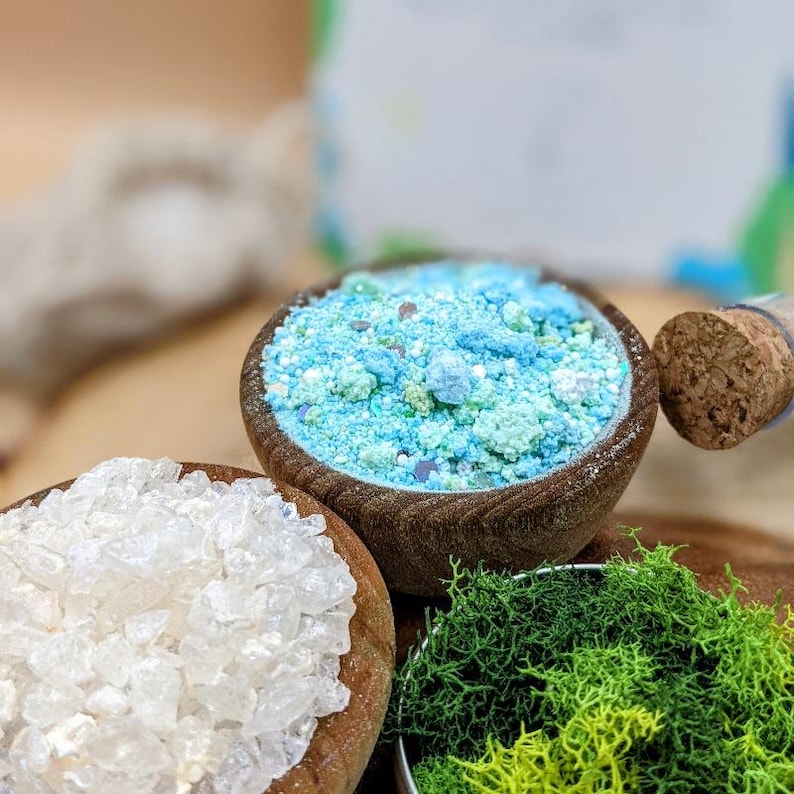 May include: Close-up of three small wooden bowls filled with colorful bath products. One bowl contains white crystals, another has blue and green granules, and the third holds green moss. A cork is visible in the background.