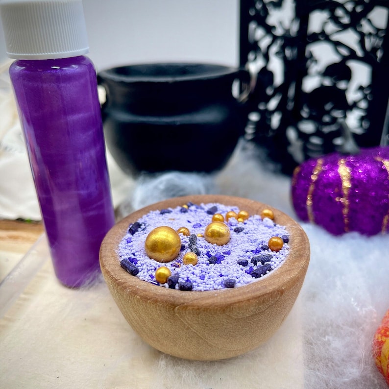 May include: A wooden bowl filled with purple and gold sprinkles. The bowl is sitting on a white surface. There is a small bottle of purple liquid and a black cauldron in the background.