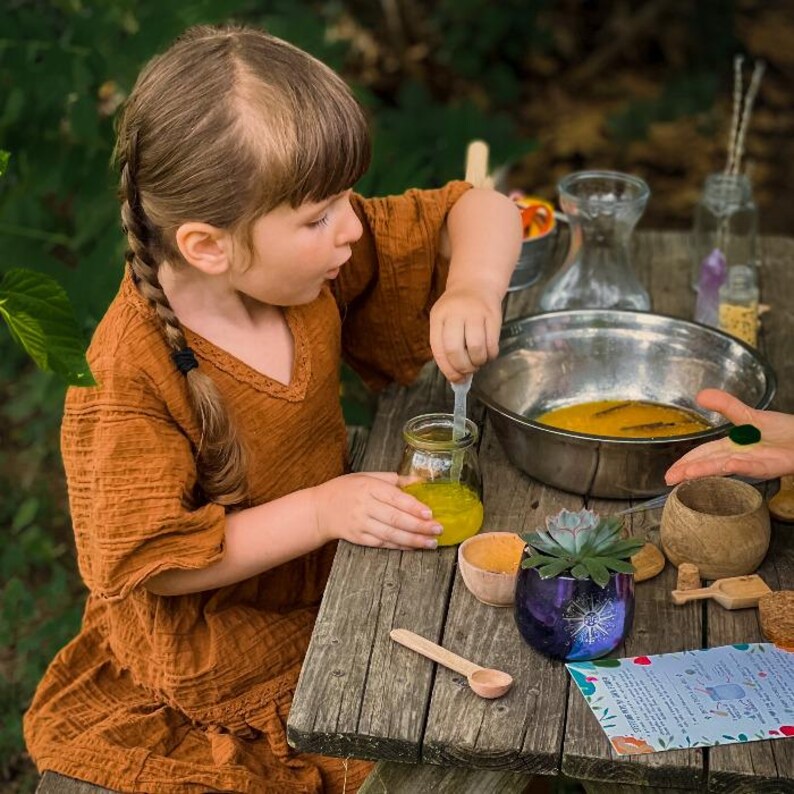 May include: A young person wearing a brown dress stirs a yellow liquid in a glass jar with a plastic spoon. The jar is on a wooden table with a small wooden spoon, a purple planter with a green succulent, and a small bowl with a yellow liquid.