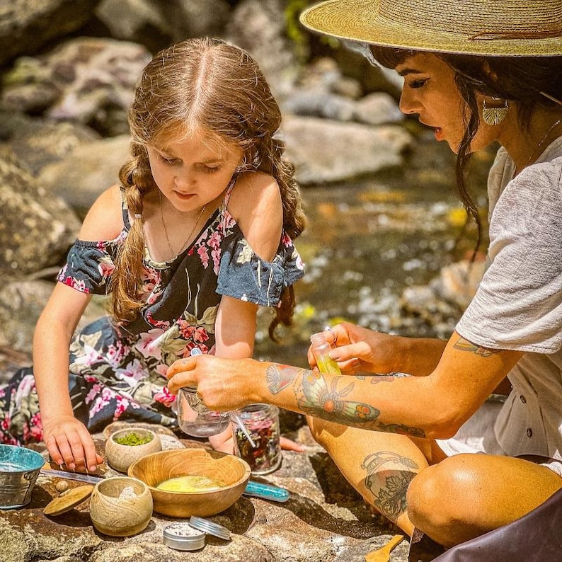 May include: A young girl and an adult are seated outdoors, surrounded by natural elements. The girl wears a floral dress, while the adult has tattoos on their arm. Various wooden bowls and small containers hold ingredients, suggesting a natural skincare or craft activity.