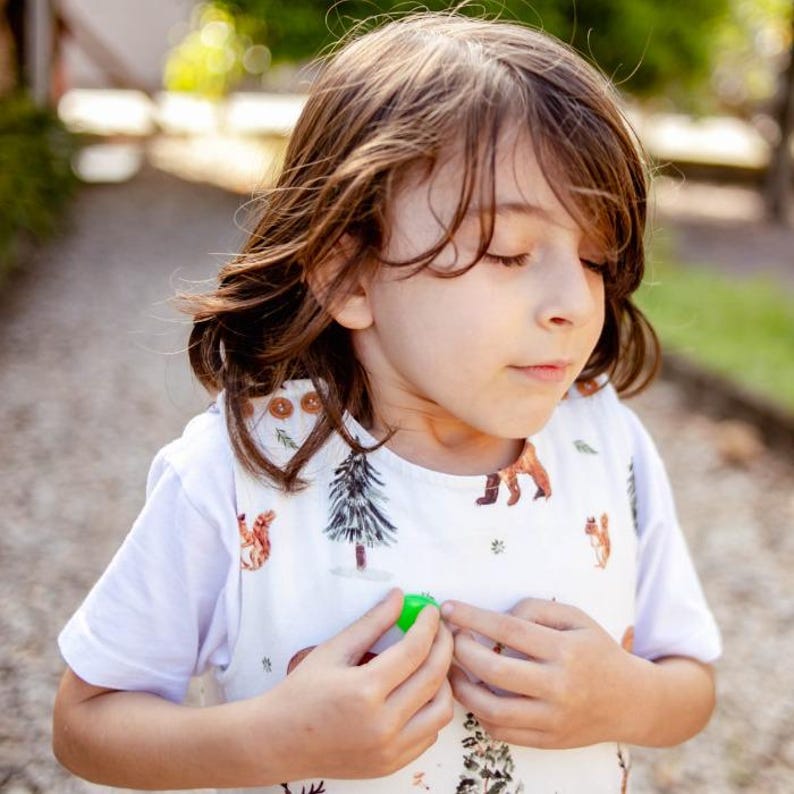 May include: A young child wearing a white short-sleeved shirt with a woodland print. The shirt features small illustrations of trees, squirrels, and deer. The child is holding a small green object in their hand.
