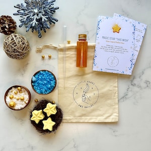 May include: A white muslin bag with a gold star and the text "Magic Star This Wish" printed on it. The bag contains a small bottle of gold glitter, a dropper, and a small bowl of blue bath salts. There are three yellow star-shaped bath bombs in a nest.