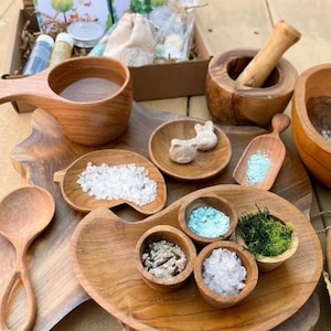 May include: A collection of wooden spa accessories, including a large cup, bowls, spoons, and a mortar and pestle. The bowls contain bath salts, herbs, and other spa ingredients. A gift box is visible in the background.