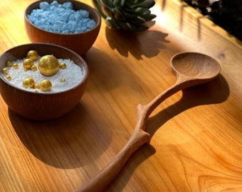 Könnte beinhalten: Ein hölzerner Servierlöffel mit einem astähnlichen Griff liegt auf einer Holzoberfläche. Zwei kleine Holzschalen enthalten weiße Granulate und goldene Kugeln sowie blaue Granulate. Im Hintergrund befindet sich eine Sukkulentenpflanze.