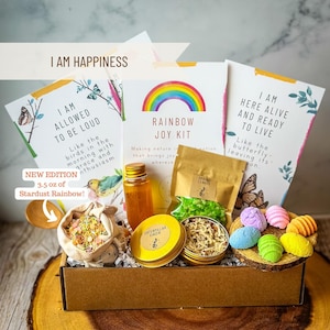 May include: A brown cardboard box filled with a rainbow joy kit. The kit includes a small bottle of liquid, a bag of green and white sprinkles, a small tin of caterpillar cheer, a bag of colorful sprinkles, and a set of colorful bath bombs. The box has a label that says "I am Happiness" and "Rainbow Joy Kit".