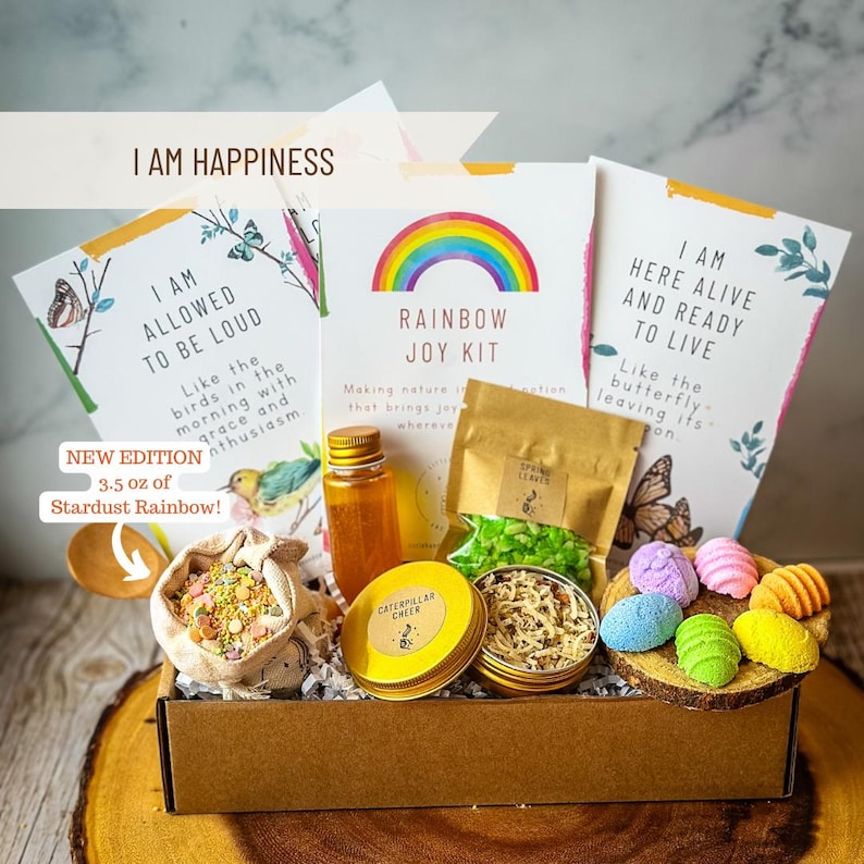 Puede incluir: Una caja de cart&oacute;n marr&oacute;n llena de un kit de alegr&iacute;a arco&iacute;ris. El kit incluye una peque&ntilde;a botella de l&iacute;quido, una bolsa de purpurina verde y blanca, una peque&ntilde;a lata de &bdquo;Caterpillar Cheer&ldquo;, una bolsa de purpurina de colores y un conjunto de bombas de ba&ntilde;o de colores. La caja tiene una etiqueta que dice &bdquo;Soy la felicidad&ldquo; y &bdquo;Rainbow Joy Kit&ldquo;.