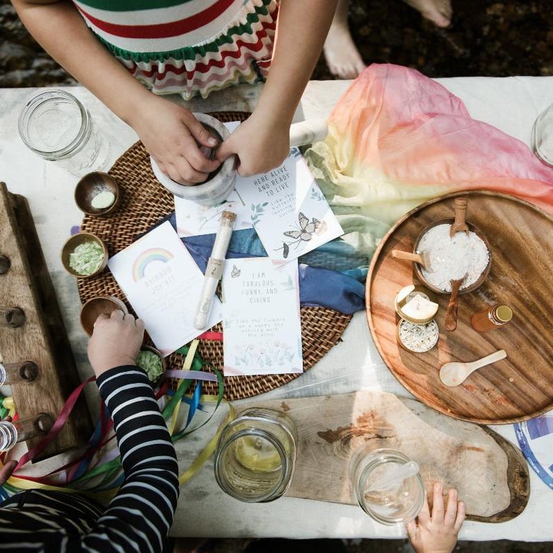Puede incluir: Una mesa preparada para una actividad infantil con tem&aacute;tica de arco&iacute;ris. La mesa est&aacute; cubierta con un mantel blanco y tiene una variedad de art&iacute;culos, incluyendo un mortero y un pil&oacute;n, tazones peque&ntilde;os, frascos y una bufanda de seda de colores arco&iacute;ris. Tambi&eacute;n hay varias tarjetas con texto, incluyendo "Here Alive and Ready to Live" y "I am Fabulous, Funny and Fiving".