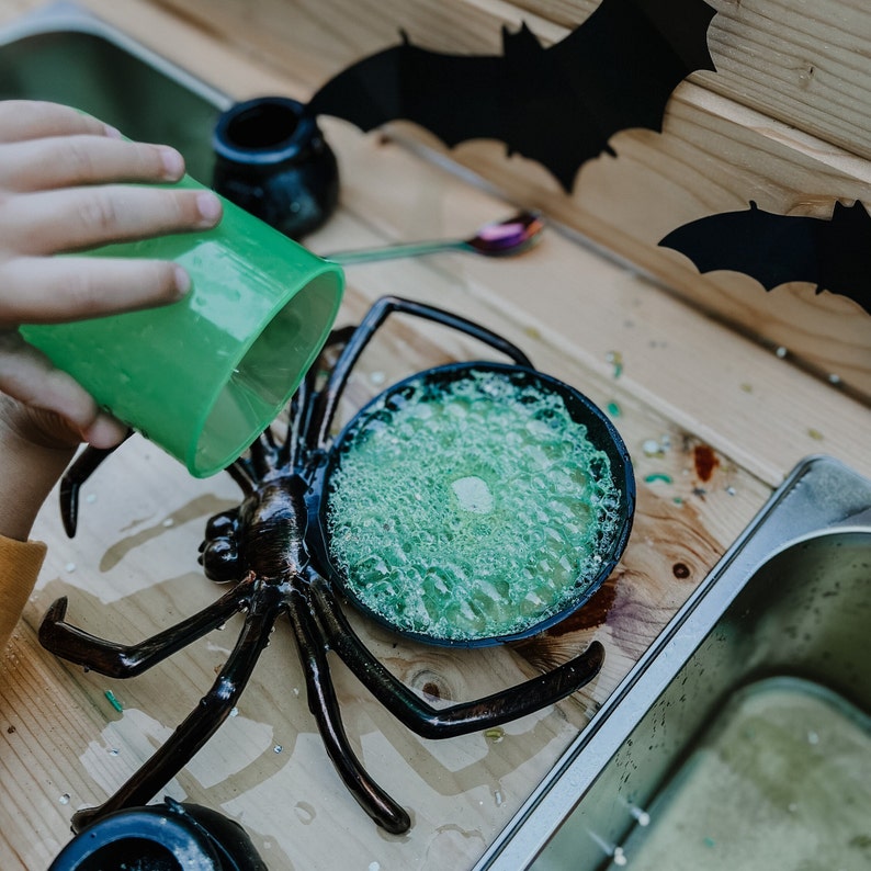 May include: A black plastic spider toy with a green slime-filled bowl on its back. The spider is on a wooden surface with a stainless steel sink in the background.