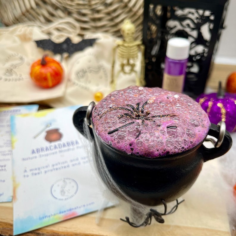 May include: A black cauldron with a pink and purple bubbling potion and a black spider on top. The cauldron is surrounded by white spider webs.