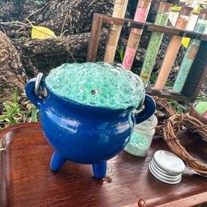 May include: A small, blue cauldron with a bubbling, green potion. The cauldron has three legs and two handles. Beside it are glass vials with colorful contents, a small jar of glitter, and a decorative wreath.