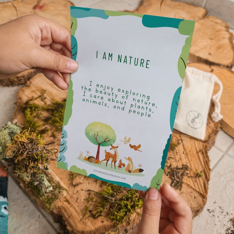 May include: A card with the text "I AM NATURE" and a nature-themed illustration. The card is held over a wooden surface with moss. A small drawstring bag with a nature-themed logo is also visible.