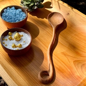 May include: A wooden serving spoon with a curved handle and a circular end, next to two small wooden bowls. One bowl contains blue crystals, and the other contains white granules and gold spheres. The items are on a wooden tray.